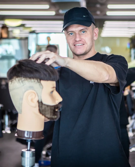 A smiling student in a black cap presenting a mannequin head with a fresh haircut inside a barbering academy. A smiling student in a black cap presenting a mannequin head with a fresh haircut inside a barbering academy.
